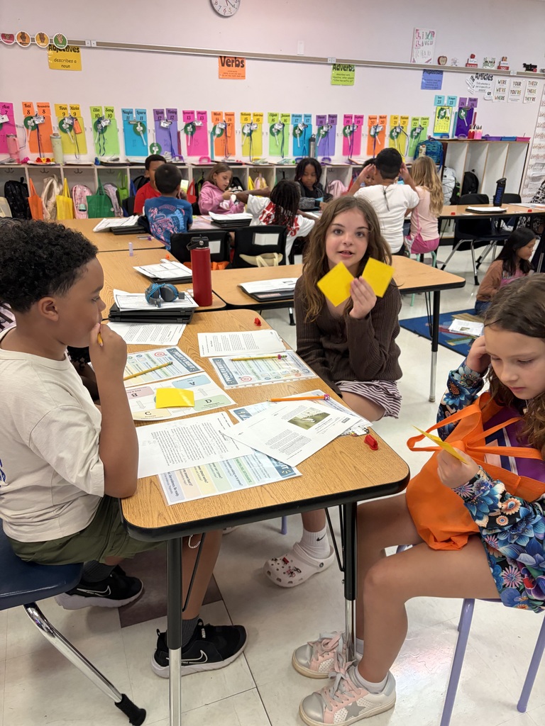 Three children in a classroom with papers on desks. A boy and two girls. Background has colorful alphabet wall.
