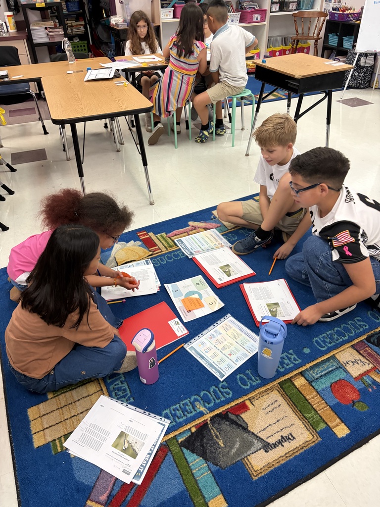Five children work on an activity with books and papers on a blue rug in a classroom.