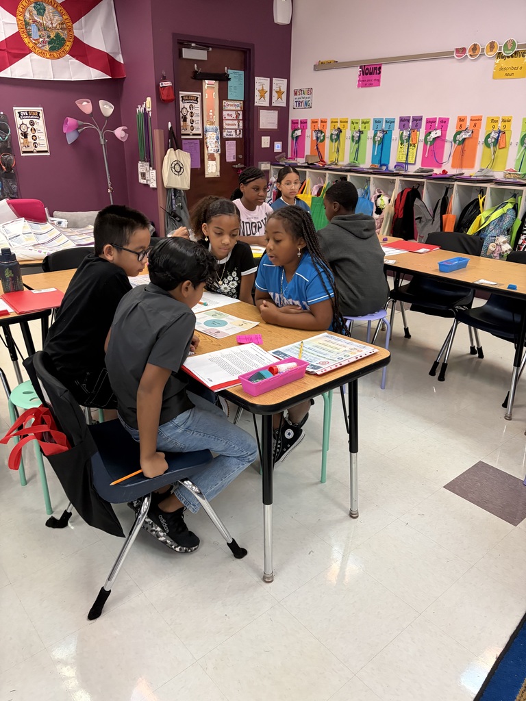 Five children sit at a table in a classroom, facing papers and pencils. A flag and a lamp are visible in the background.