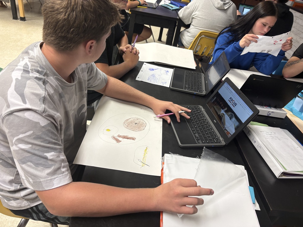 A student uses a laptop on a table in a classroom; a student behind him reads a paper.