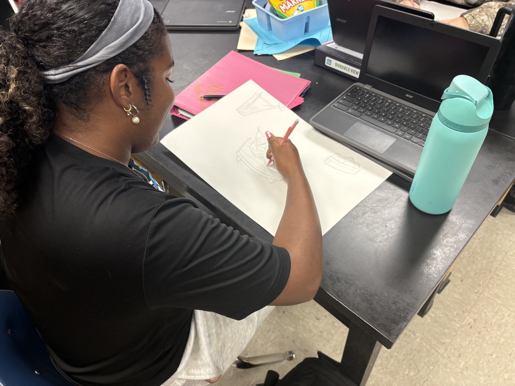Person sitting at a desk, drawing on paper with a pencil. A laptop, a blue water bottle, and folders are on the desk.