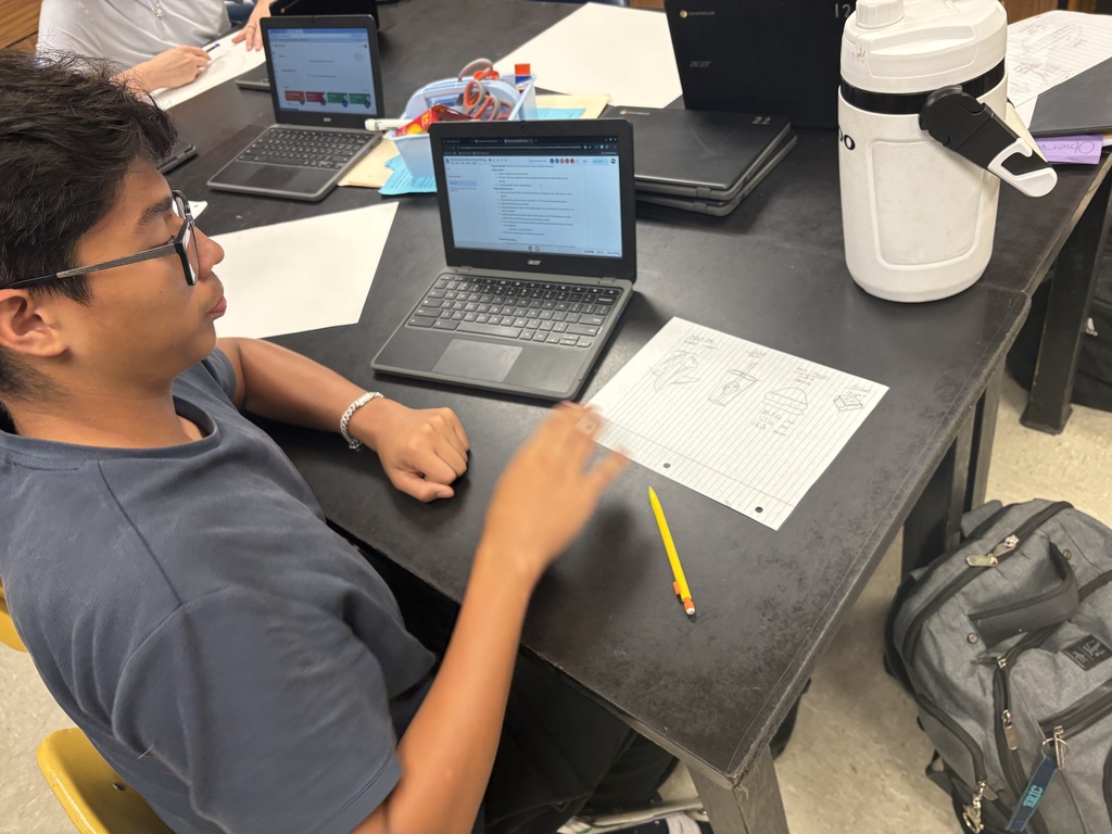 A person wearing glasses and a gray shirt sits at a table, gesturing with a pencil while a laptop is open.
