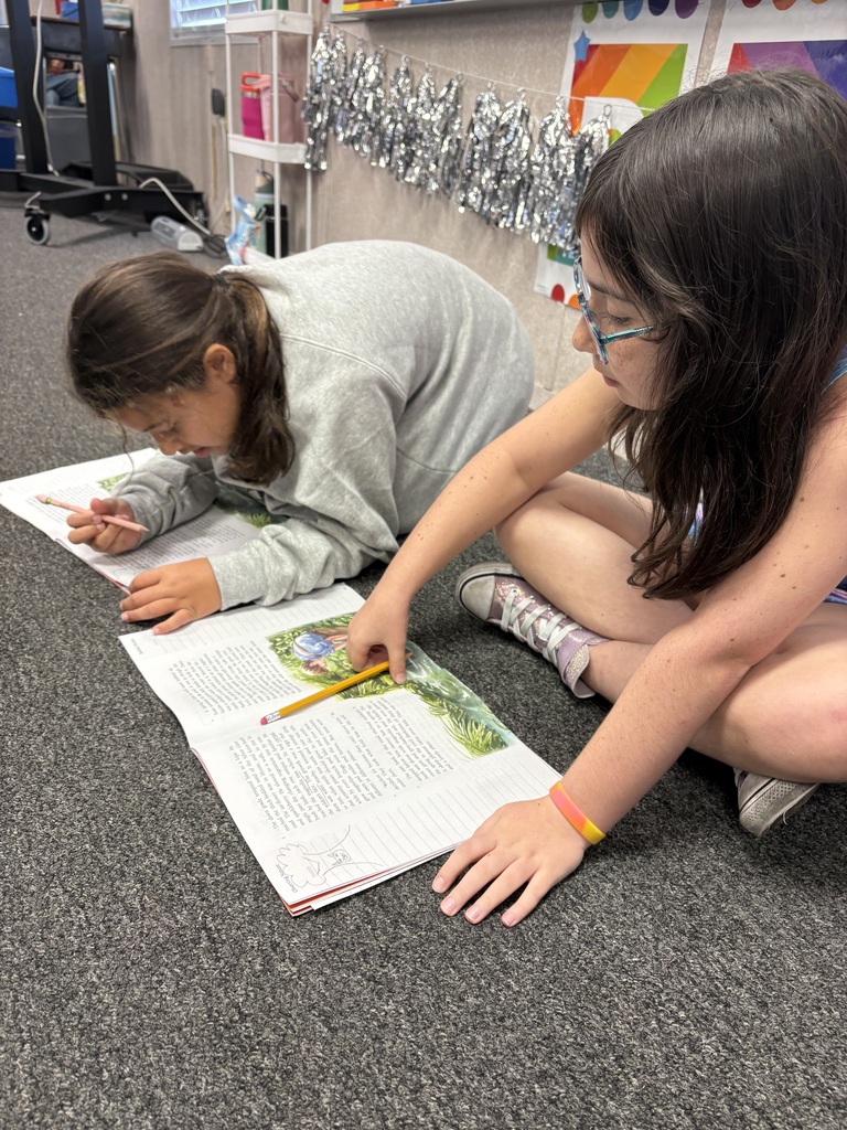Two girls on the floor, one lying down and the other sitting. Both are reading from a book.