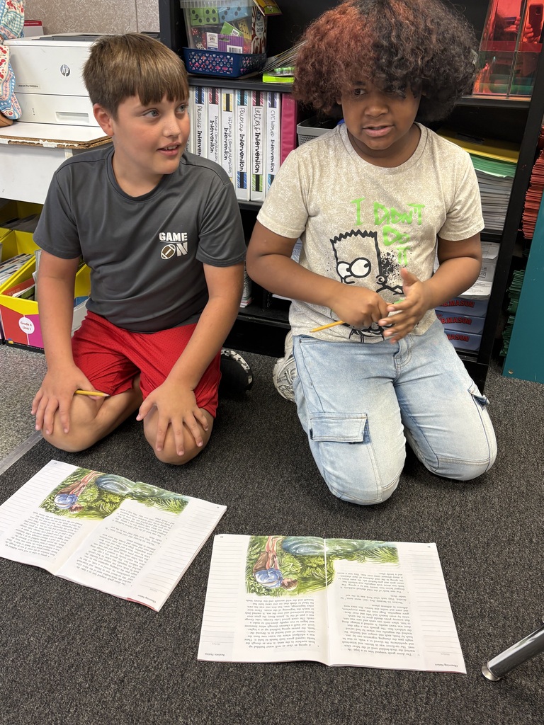 Two children sitting on the floor, one holding a pencil, both reading books on the carpet.