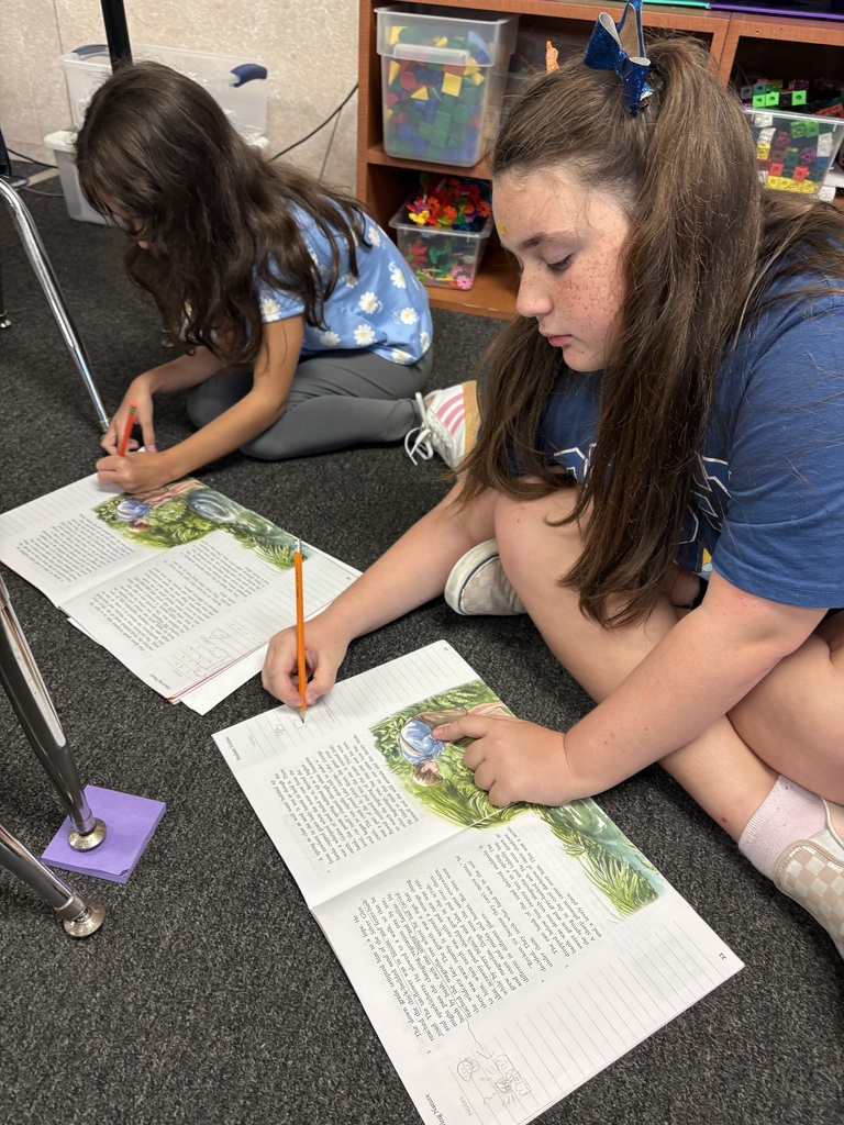 Two girls seated on the floor, writing in open books. One has a pencil in her hand.