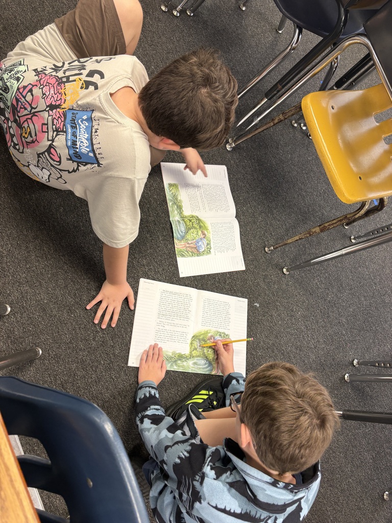 Two kids are sitting on the floor and pointing at pages of books. One kid is wearing a floral shirt.