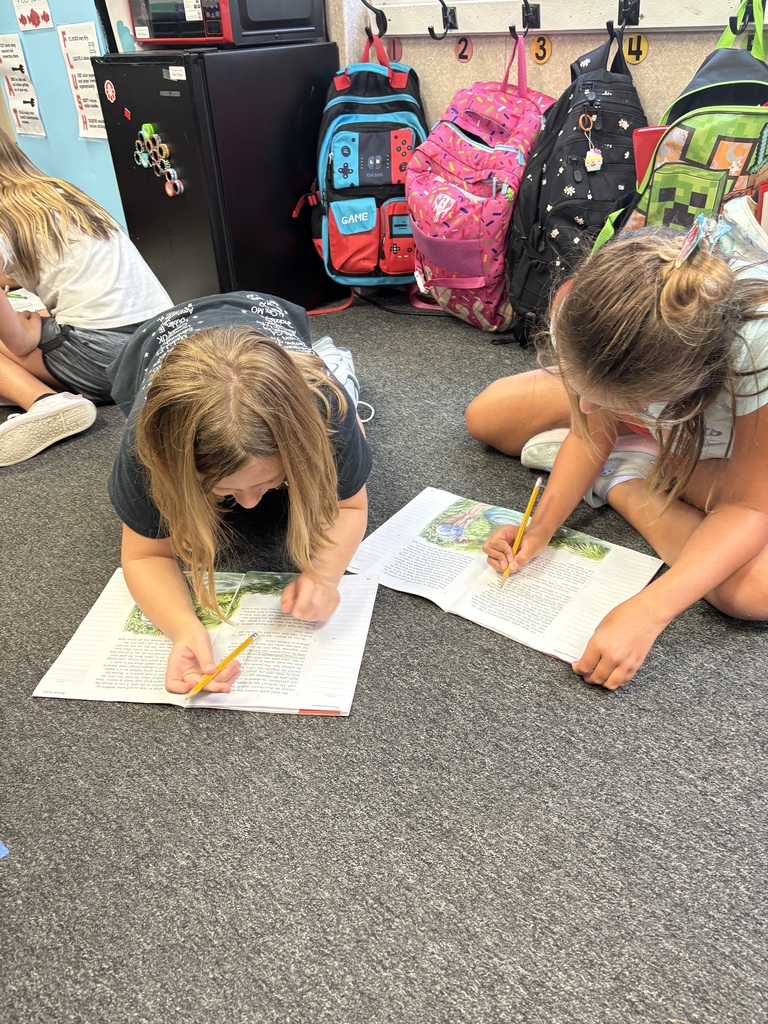 Two girls sitting on the floor, each with a book, are writing. Backpacks are in the background.