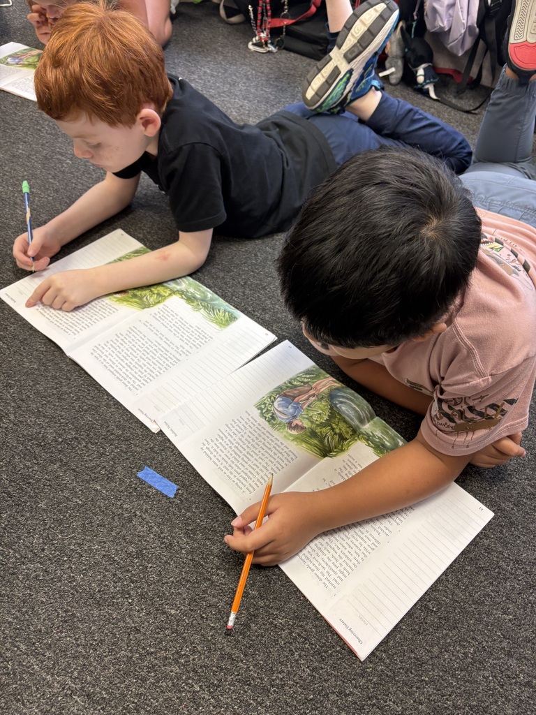 Two children work on papers lying on the floor, one with a pencil and the other with a pen.