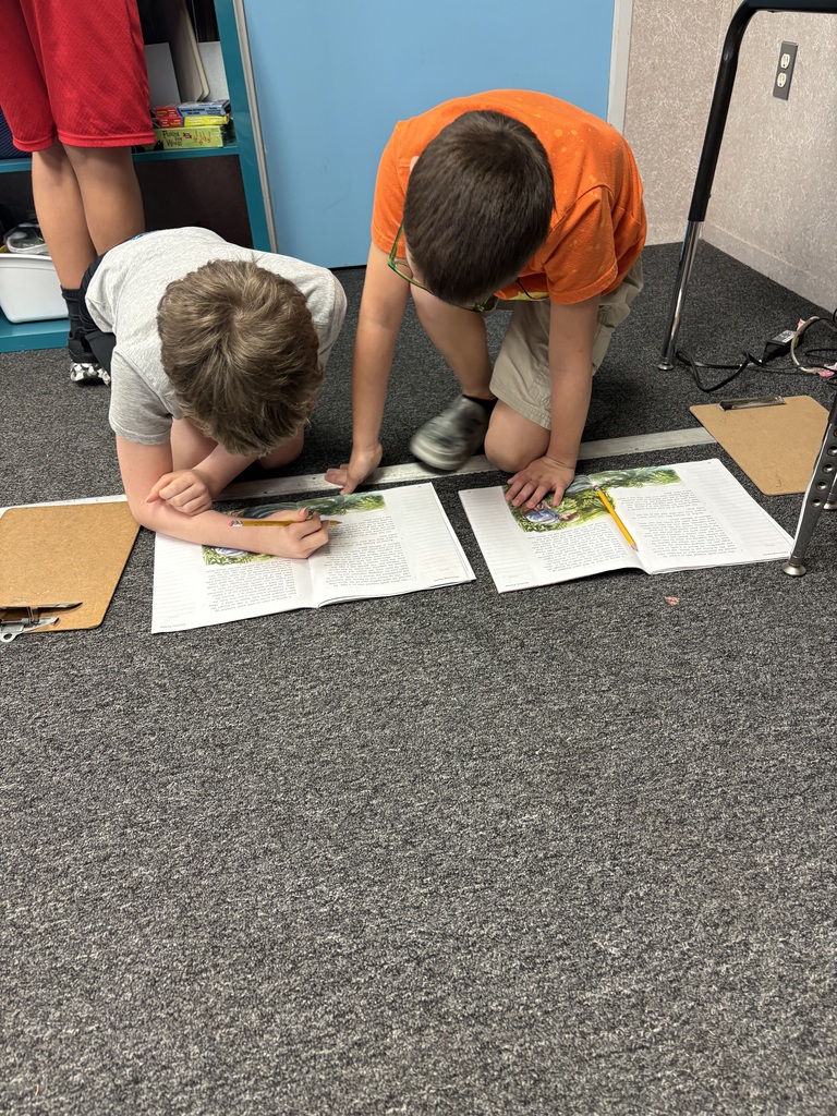 Two children on the floor, working on papers. One wearing a red shirt, the other in an orange shirt.