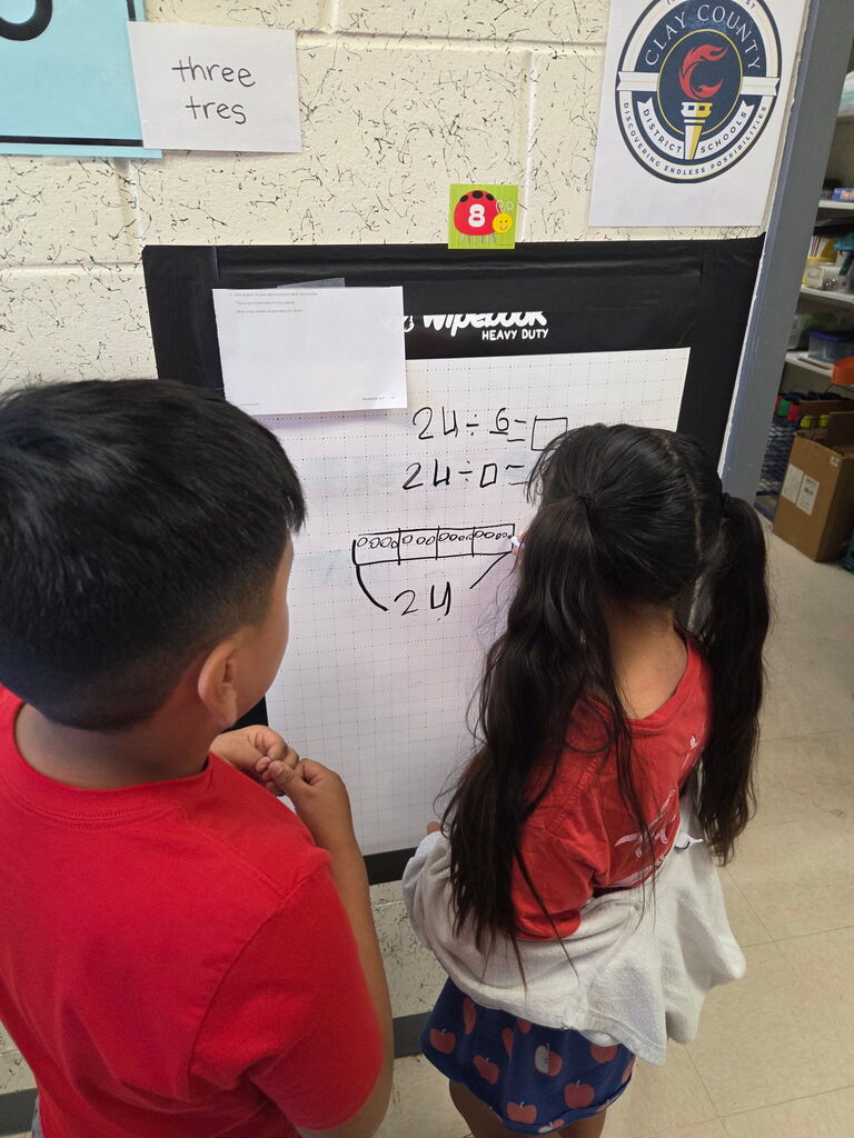 Two children, a boy and a girl, look at a whiteboard with addition problems. The boy is in red; the girl is in red and blue.
