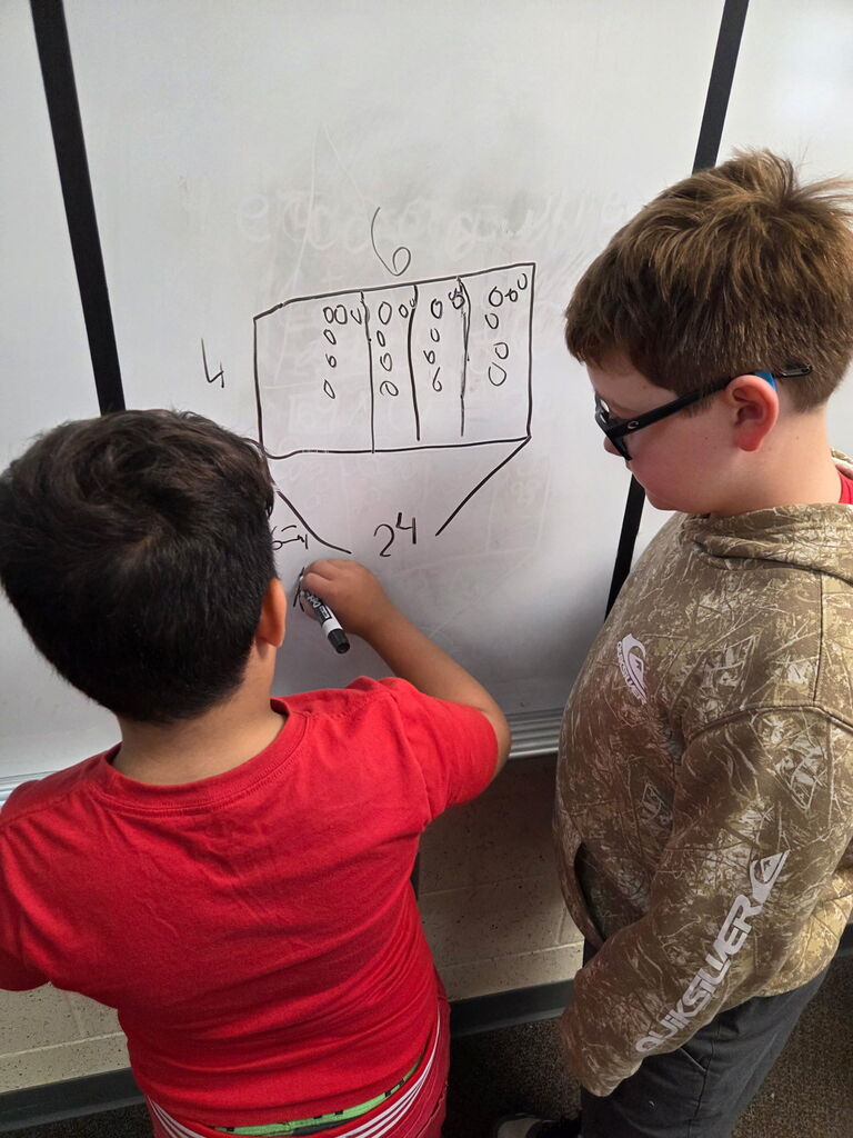 Two boys in a classroom, one writing on a whiteboard with a marker, the other wearing glasses.