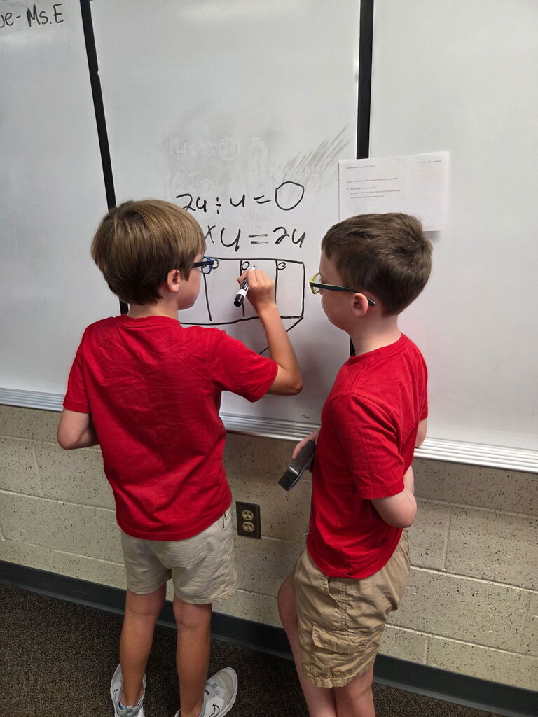 Two children in red shirts and glasses write on a whiteboard. One points to the right side.