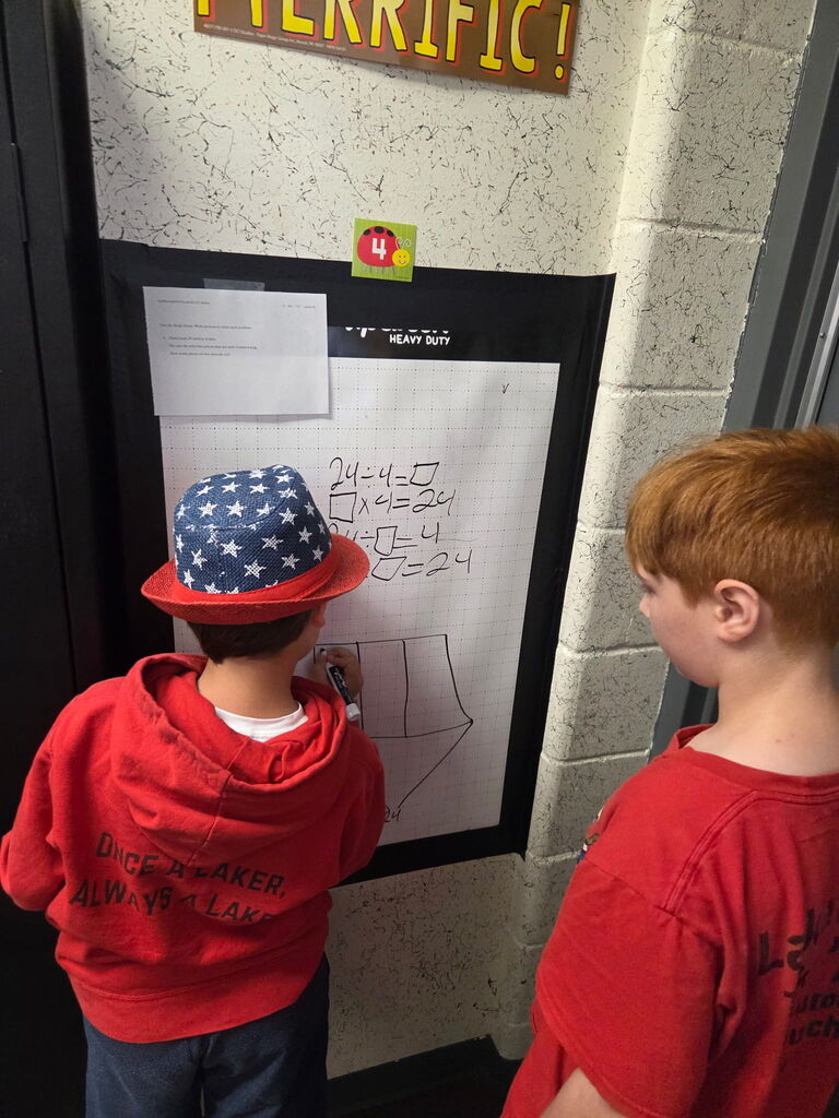 Two young boys, one in a red hat, stand near a wall with a paper sign and drawing.