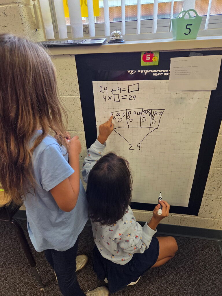 Two children drawing on a whiteboard. One child kneels, holding a marker. A third person stands.