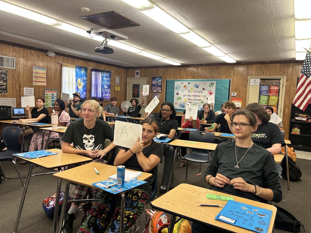 A classroom with desks, chairs, and a projector. Students sit with papers and markers, one holding a sign. A flag is in the background.