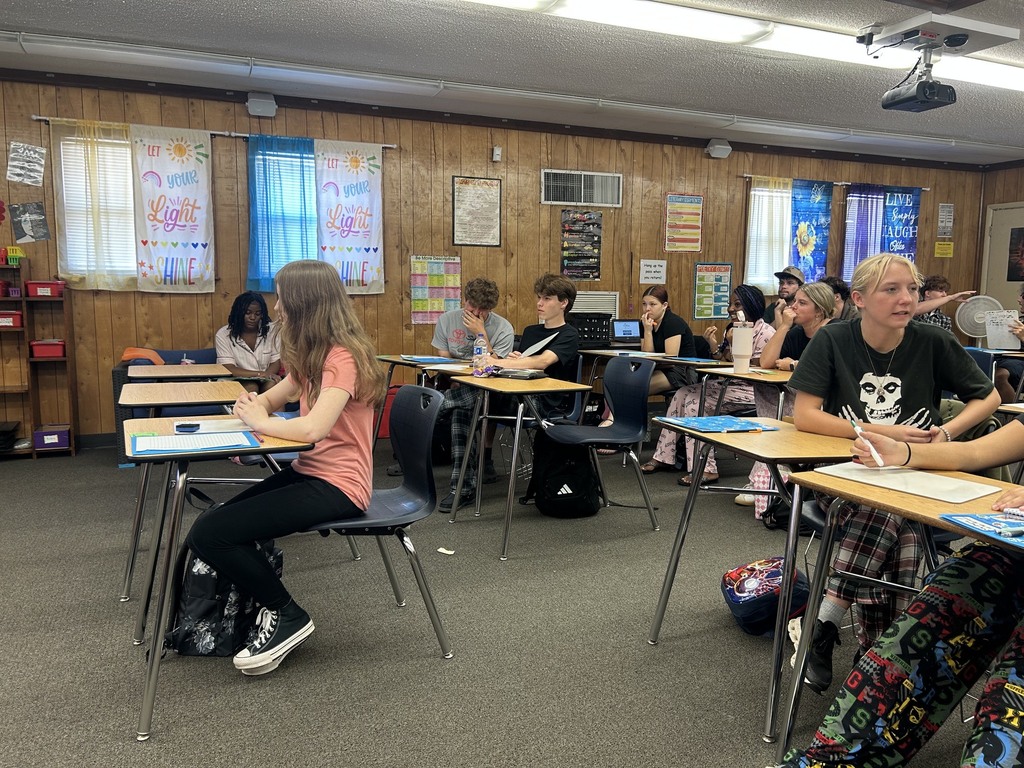An interior of a classroom with students seated at desks. Walls are wood-paneled, and a projector is mounted on the ceiling.
