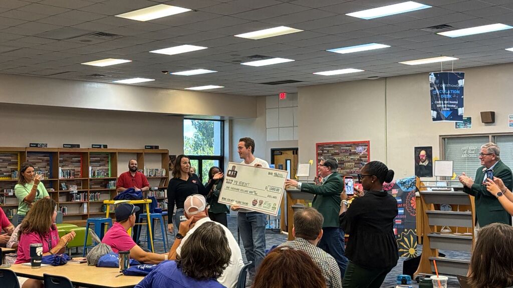 A room with tables and chairs; a group of people stand around a man holding a large check.
