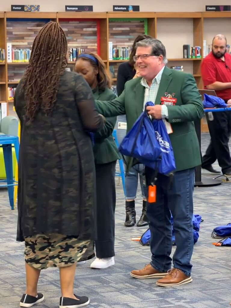 People interact in a library. A man in a green blazer holds a blue bag and smiles.