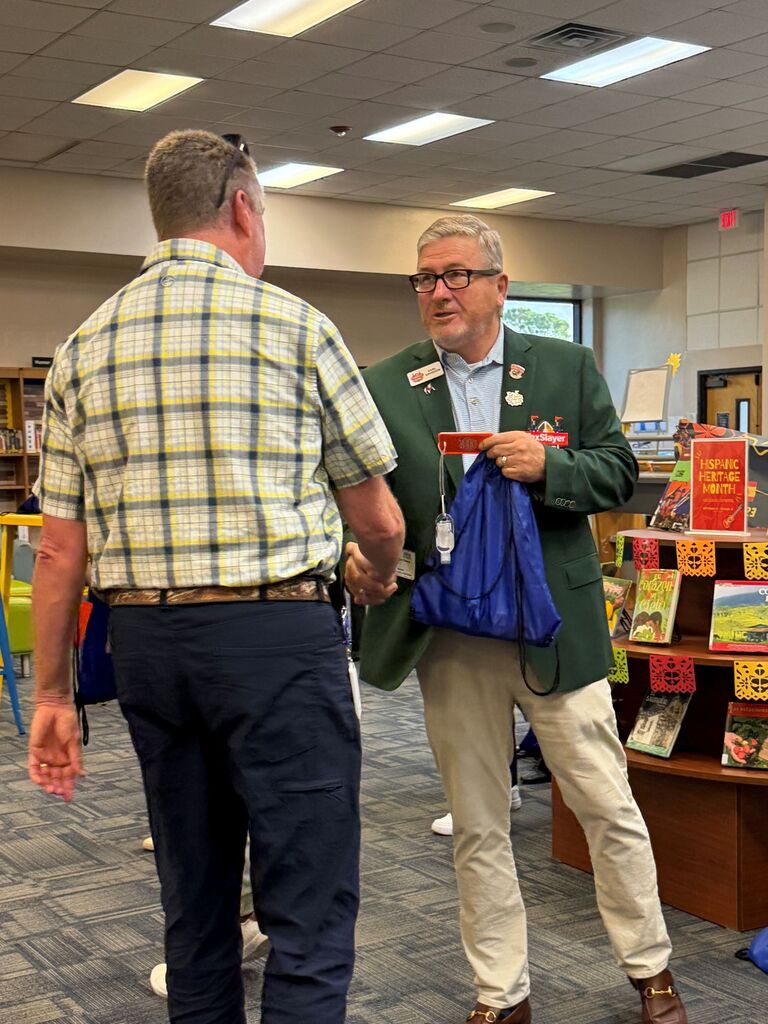 Two men in a library, one in a green jacket with a badge, talking. Behind them, a shelf with books.