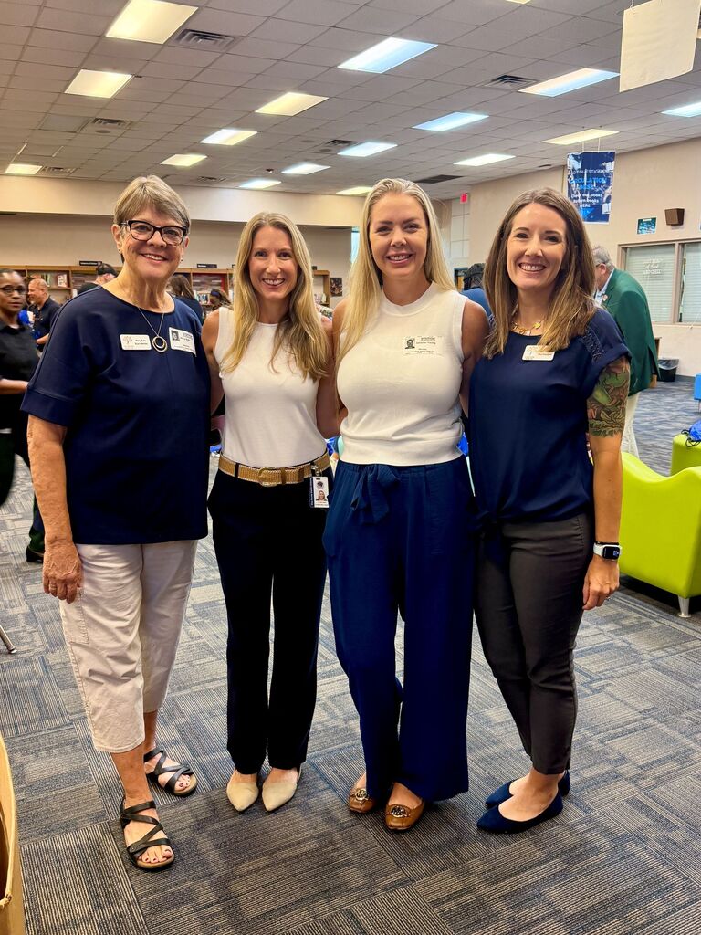 Four women stand in a room with a carpeted floor and ceiling lights. They wear blue and white shirts and pants. One has a belt.