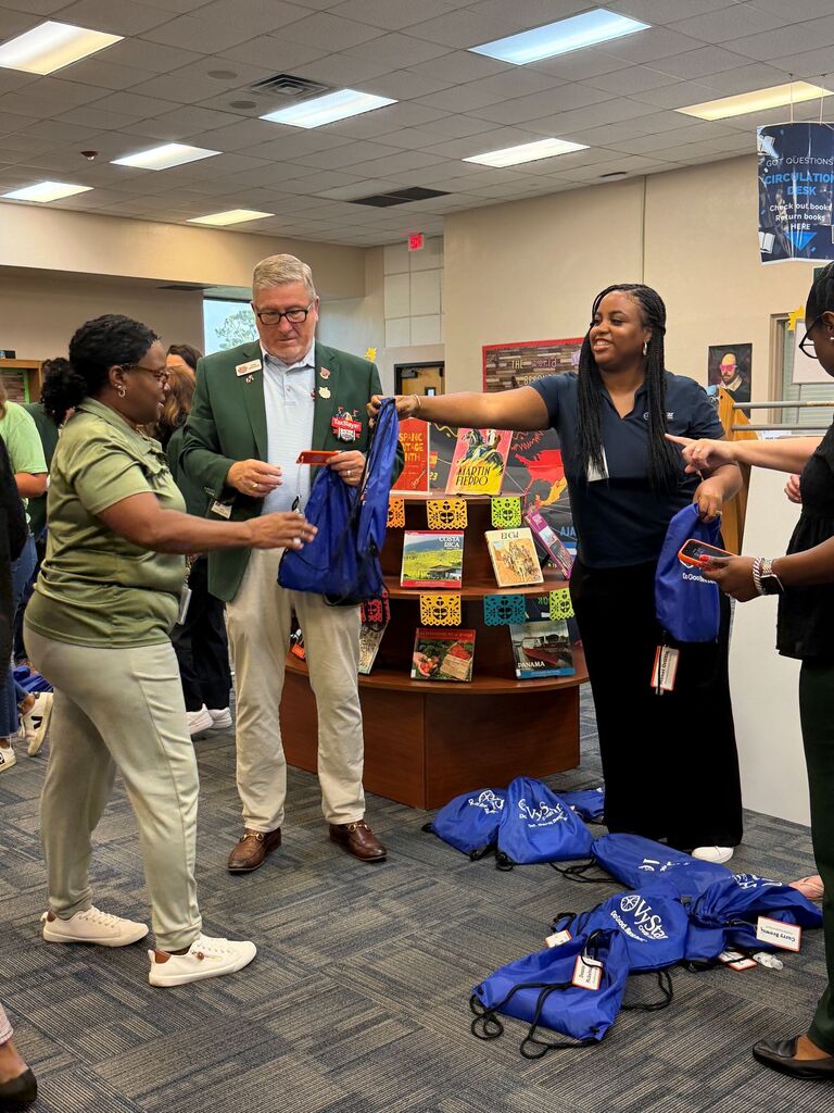 Four adults inside a room, one man hands out bags to three women, blue bags on floor.