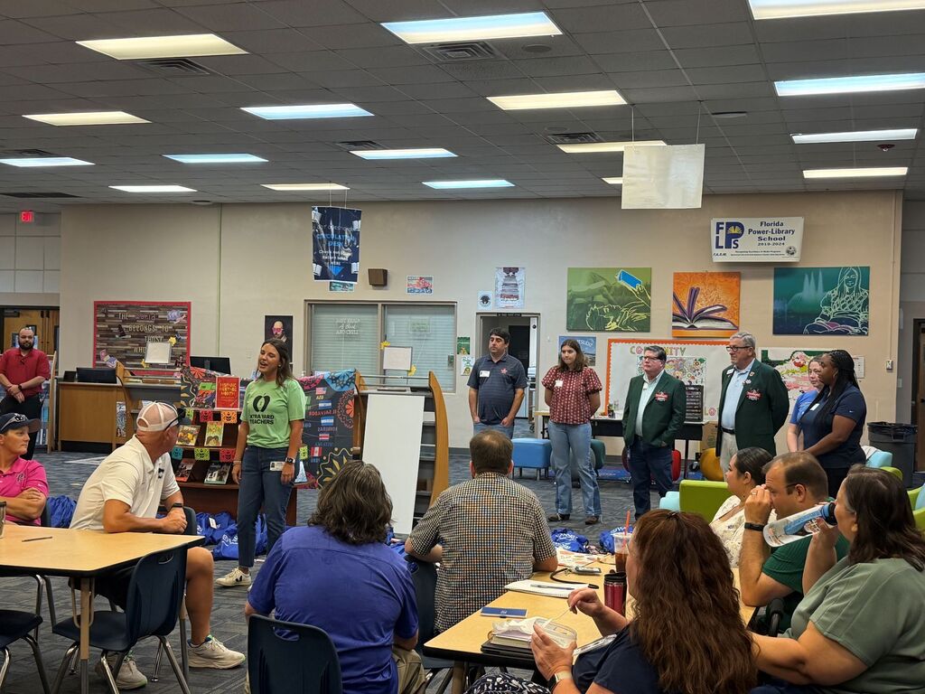 A group of people stand and sit around tables in a room with posters on the wall.