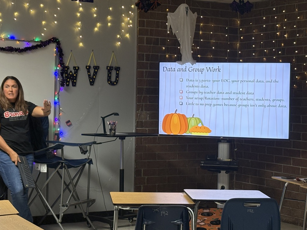 Woman standing in front of a screen with a presentation. Desks and chairs are arranged in front of her.