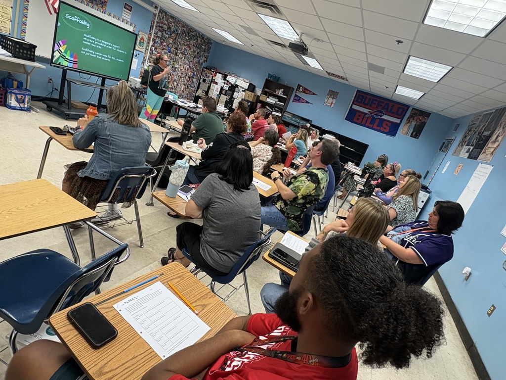 A classroom with blue walls, ceiling lights, and a teacher presenting at the front. Students seated at desks, facing the teacher.