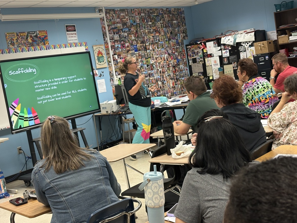 A teacher standing before a large screen presents to a class of students seated at desks in a classroom with blue walls.