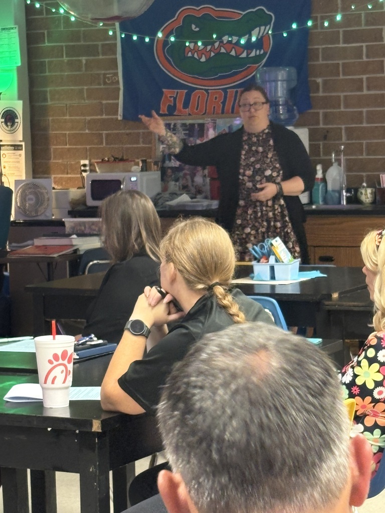 A woman stands at a desk in front of a brick wall with a banner. Several people sit at tables, listening.