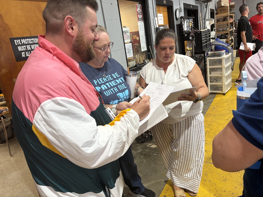 A man in a multicolored jacket is reviewing a document with a woman in a white top. They are surrounded by a few people in a room with yellow flooring.