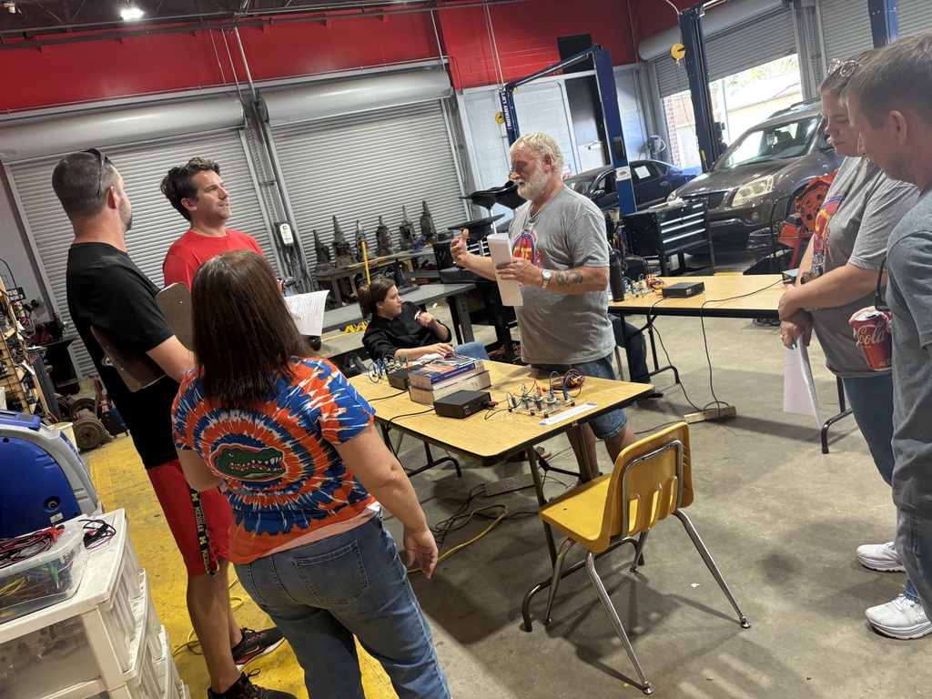 Several people stand in a workshop with tables and tools, engaged in conversation. Bright lighting illuminates the area.