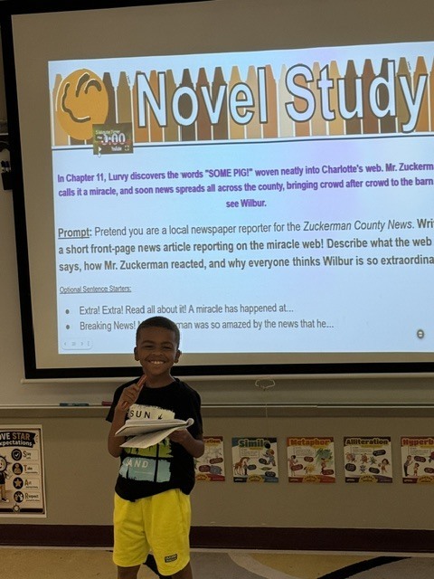A child stands in a classroom, holding a paper. Behind him, a screen shows a slide with text.