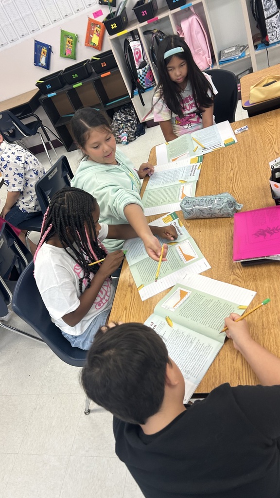 Four children sit at a table with papers, pencils, and books. They are working together