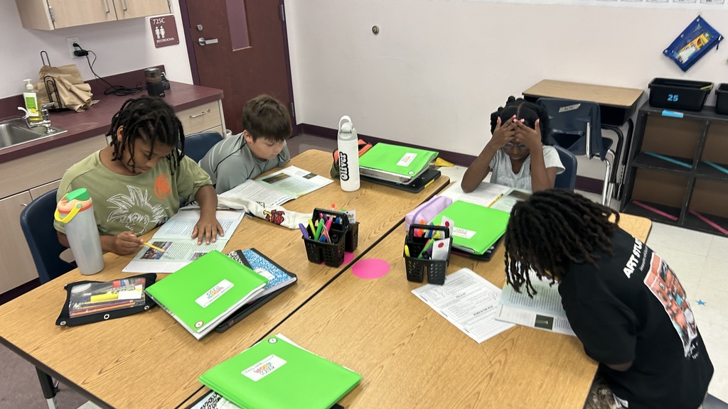 Four children in a classroom, seated at a desk, reading together