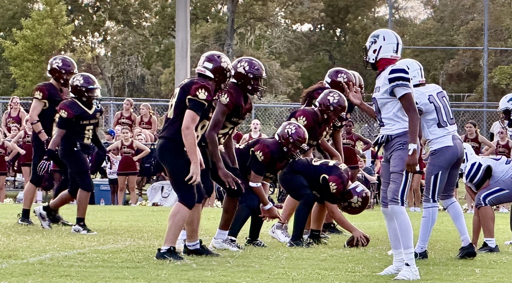 Team wearing maroon helmets plays football on a field. Team wearing white helmets in huddle. Spectators watch from the stands.
