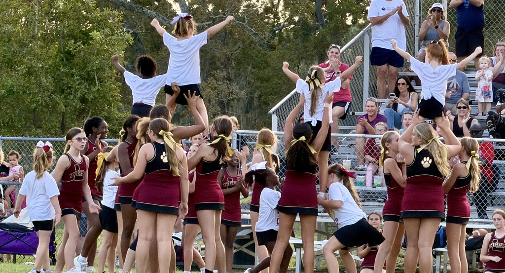 A cheerleading team performs a stunt on a field, surrounded by a crowd of people watching from the bleachers.
