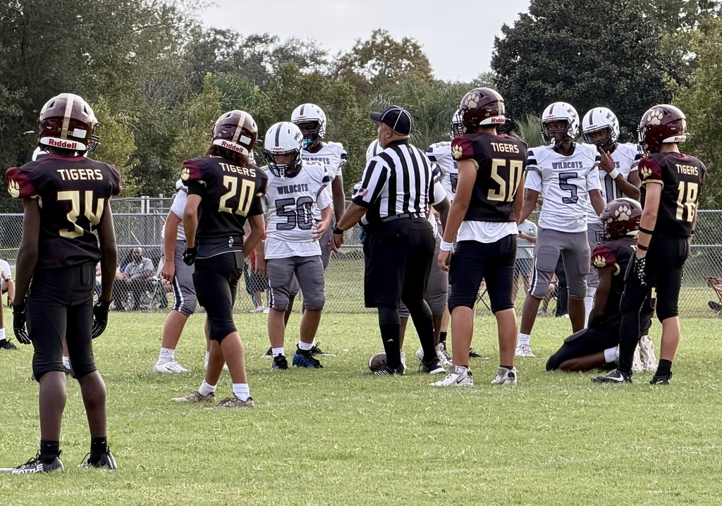A group of football players wearing helmets and jerseys huddle together on a grassy field. The background includes trees.
