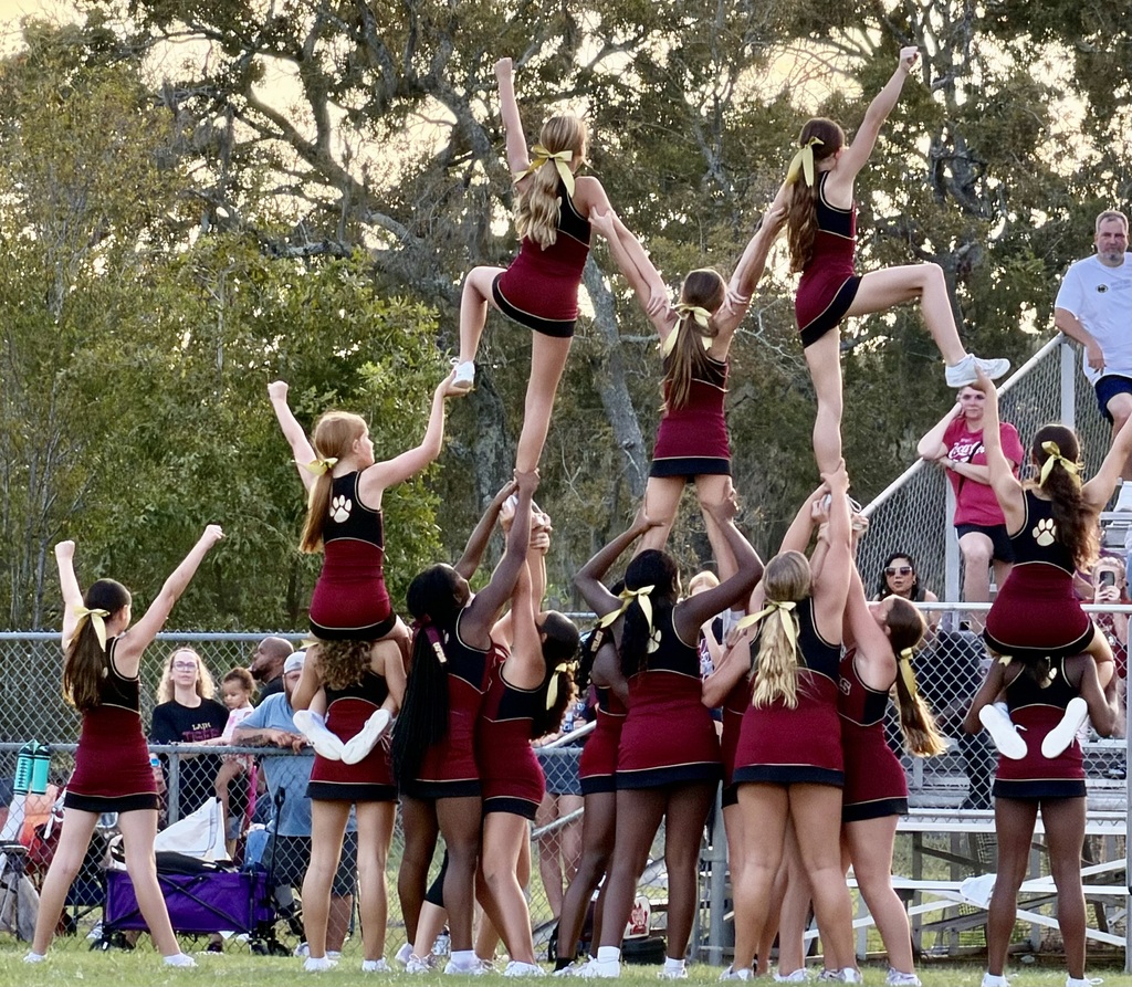 Cheerleaders performing a pyramid stunt, with others watching in the background. Trees and bleachers visible.