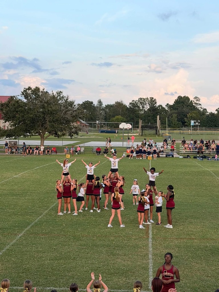 Group of cheerleaders forming a pyramid, spectators watching in the background with trees and a setting sun.