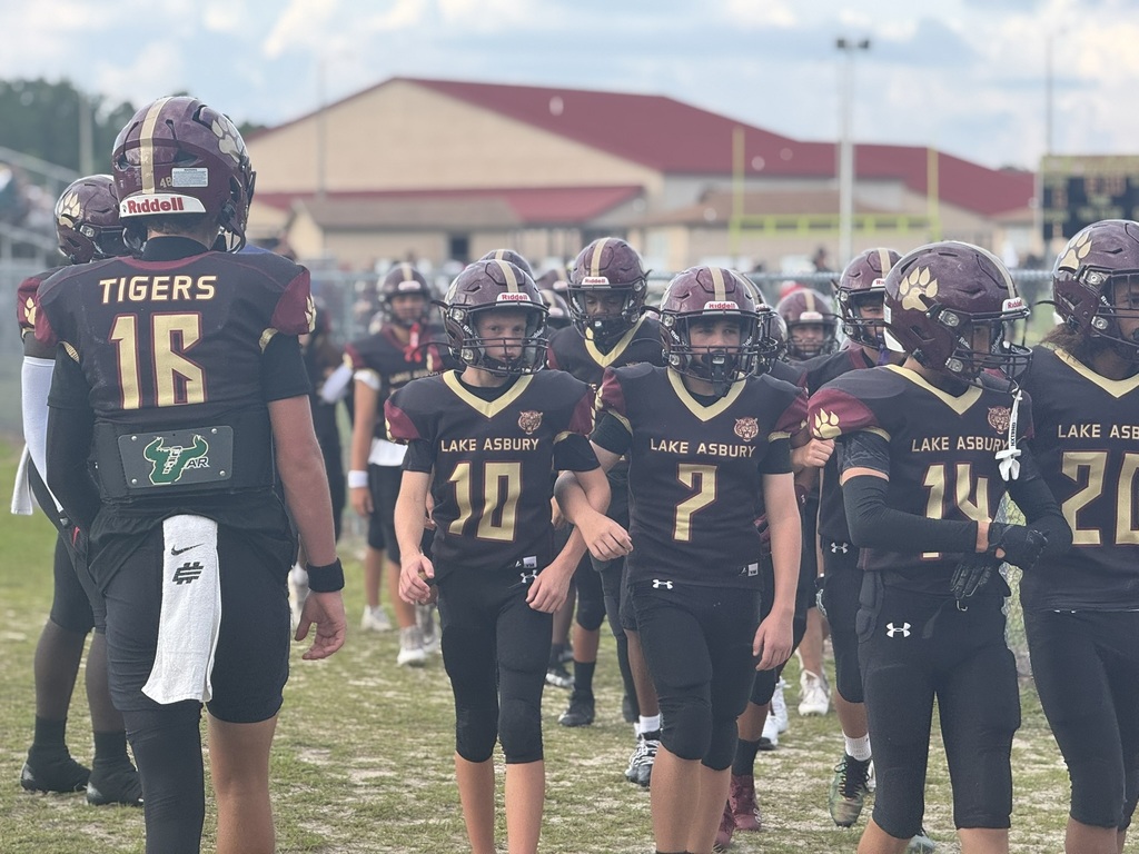 American football players in black uniforms with gold numbers walk onto a field with a building in the background.