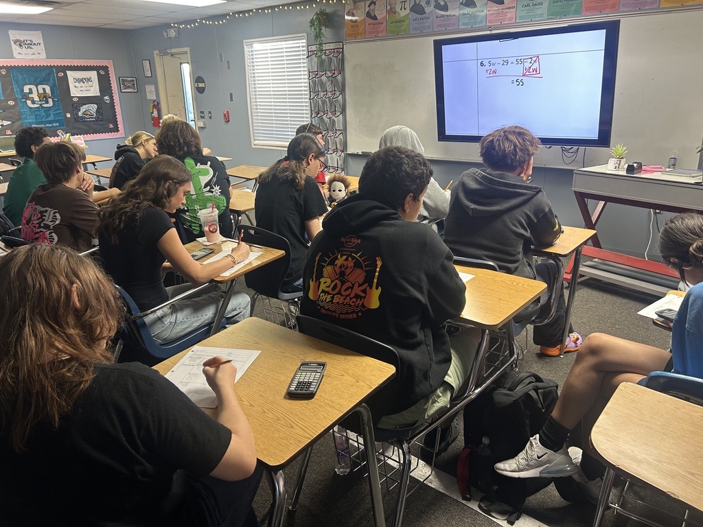 Classroom with desks, students seated, and a teacher at the front with a large screen and whiteboard.