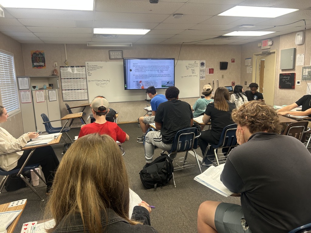 A classroom with students seated at desks. A projector screen displays content. Students and a teacher interact.