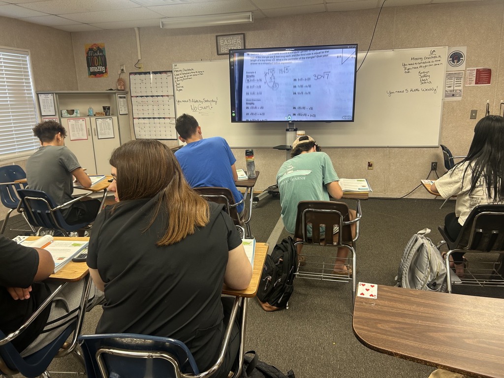A classroom with a flatscreen monitor on the wall. Students are sitting on chairs and writing.