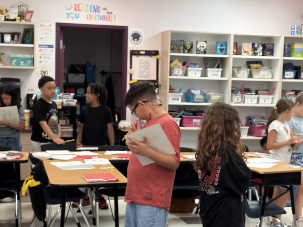 Children in a classroom stand around tables, with some holding papers. 