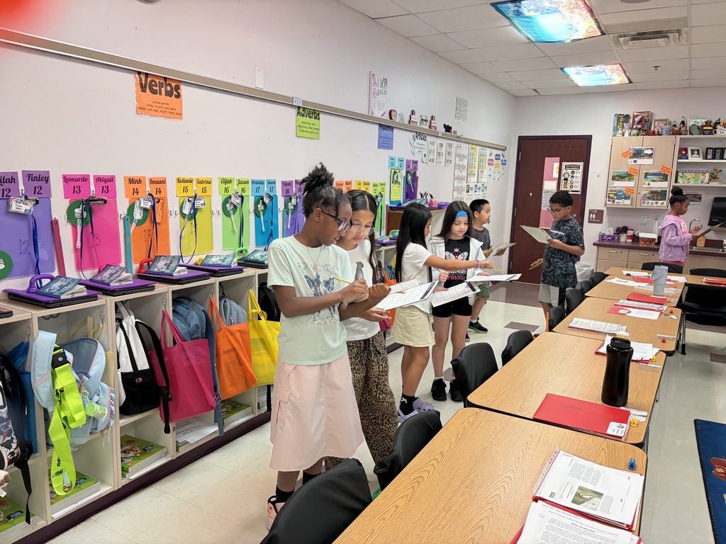 Five children stand in a classroom, reading papers. 