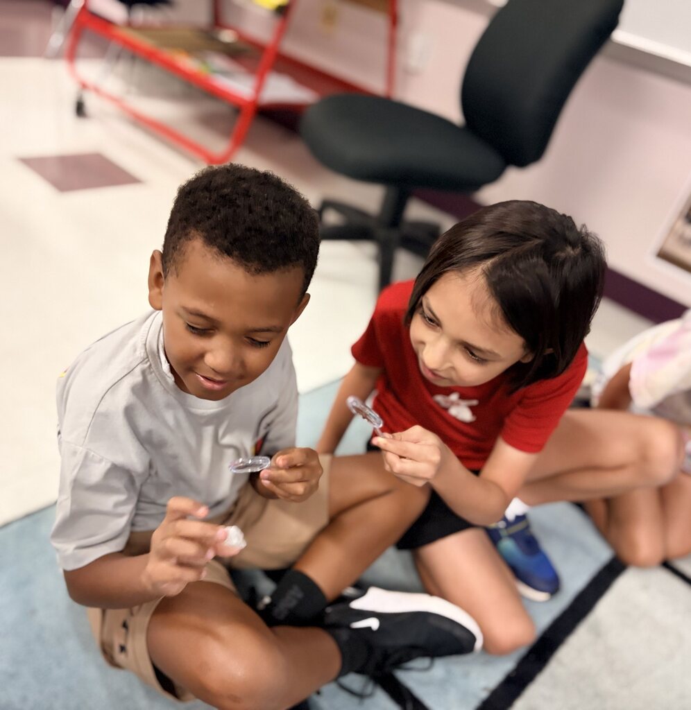Two kids sit on a rug, smiling. One holds a small rock