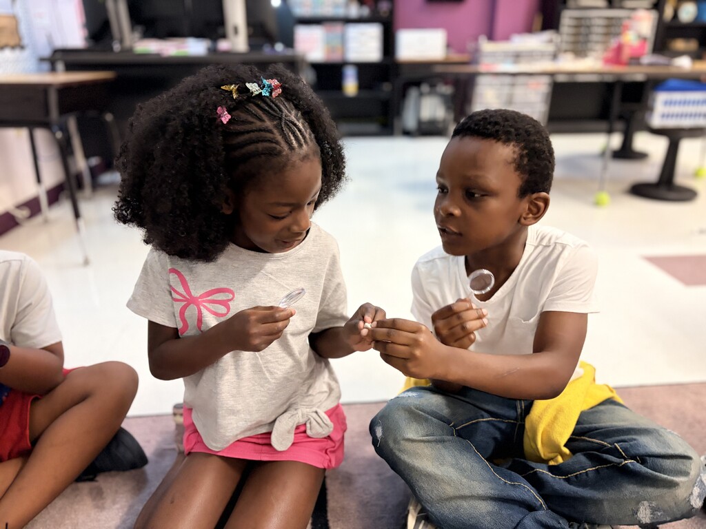 A boy and girl are sitting on the floor. The girl is holding a rock and the boy holds a magnifying glass.
