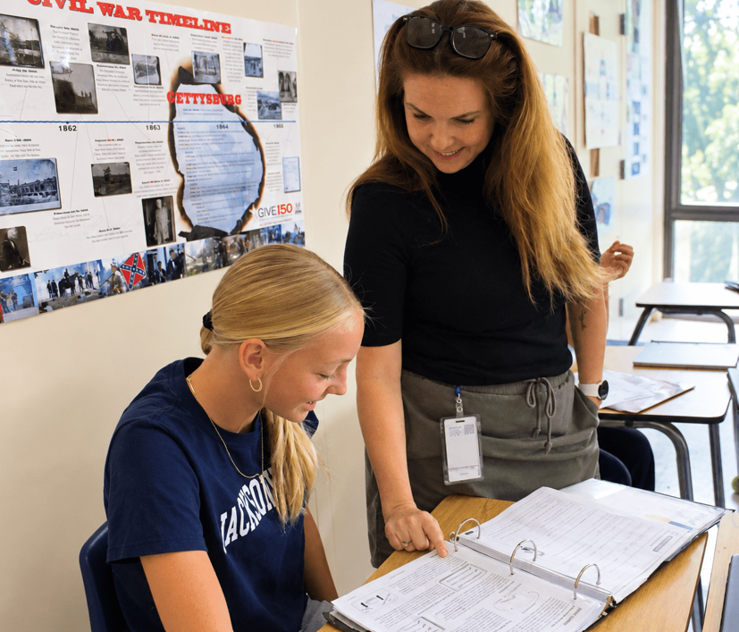 Two women in a classroom, one studying a binder while the other stands behind her. A Civil War timeline is on the wall.