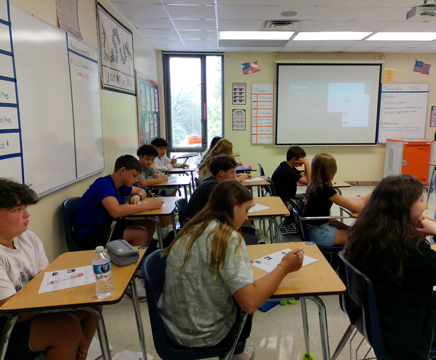 Students in a classroom, sitting at desks with notebooks and pens, and a projector screen at the front.